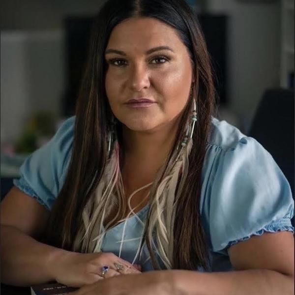 Brandi is wearing a blue shirt and sits with her arms on a table in front of her