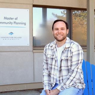 A man wearing a beige and white plaid jacket over a white t-shirt is sitting on the arm of blue Adirondack chair in front of a door and window to a building