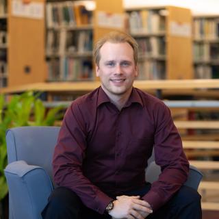 Chris Shanks sits on a blue chair in a library.