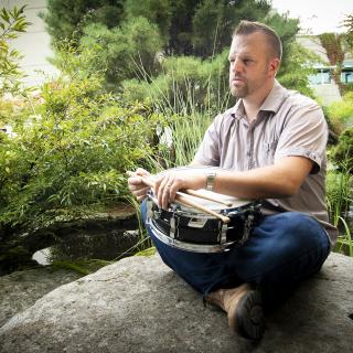 VIU Music Department Instructor Hans Verhoeven sits on a rock near a pond holding a drum.