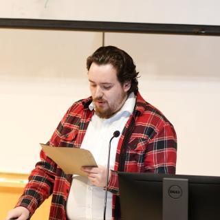 A student reads off of a piece of paper at the front of the classroom.