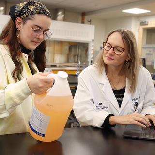 Lily Eggert and Dr. Alexandra Weissfloch look at a bottle of cleaning product, which is an amber colour, while in a VIU chemistry lab. Both wear lab coats.