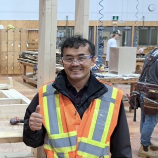 Ezat Haidary wering a reflective vests and safety goggles in the carpentry shop, giving a thumbs up and smiling at the camera.