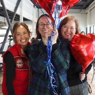 Three women smile, one holding a bunch of balloons, another holding a heart balloon near her face