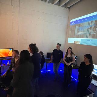 Students gather around three screens on a desk with a projection of an image on the wall behind them.