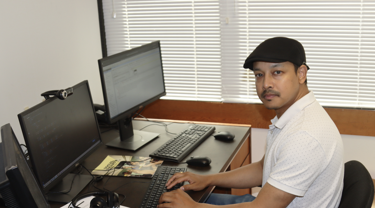 Ajay Shrestha sitting in his office, looking at the camera and in front of a keyboard and three computer screens