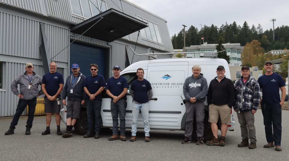 The facilities team at VIU standing outside in front of a work van on a cloudy day