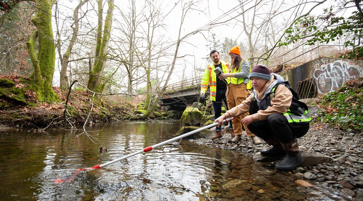 Three people next to a stream, one holds a sampling pole while the others look on