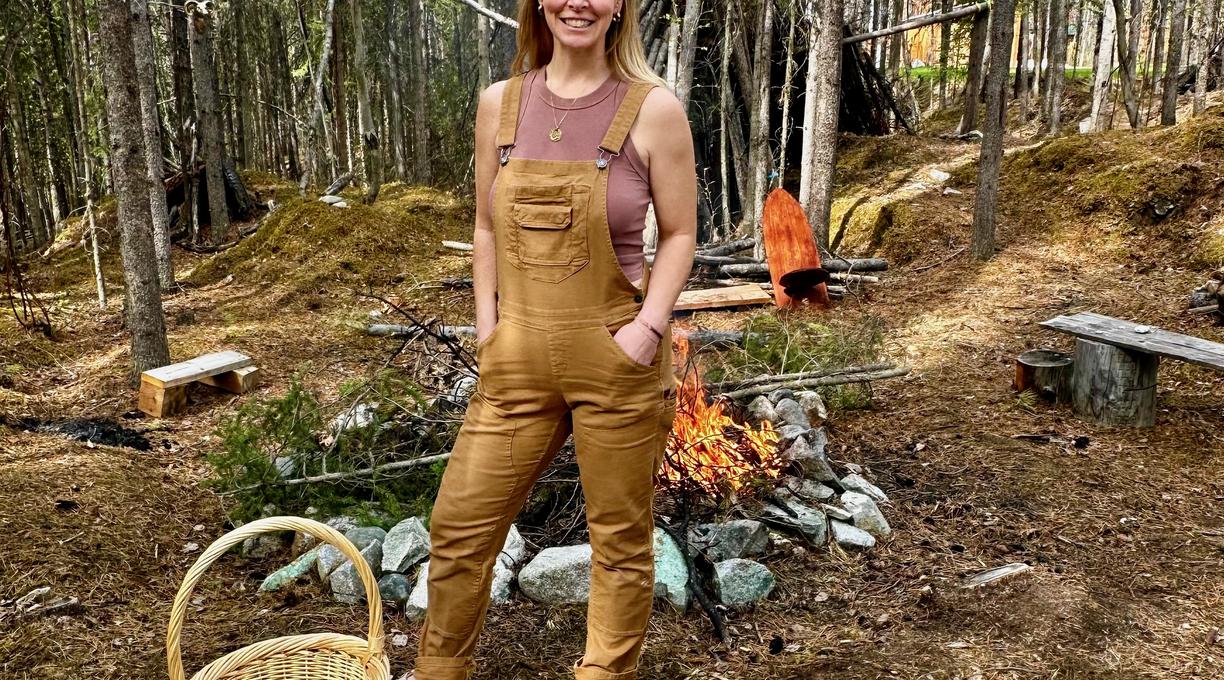 A woman wearing tan overalls is standing in a clearing in the forest in front of a campfire. 