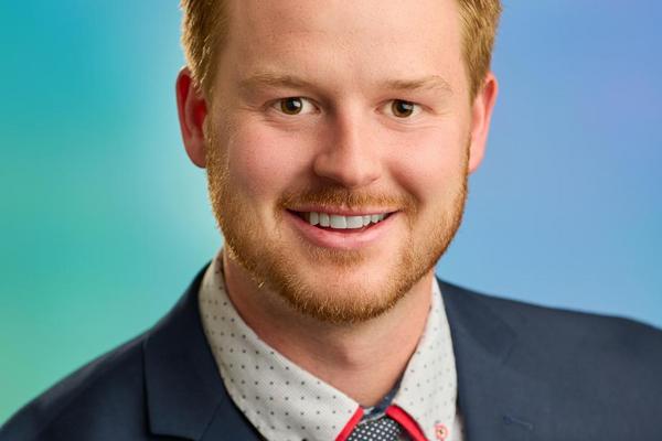 Mitch Turko wearing a suit and tie, smiling at the camera with a blue and green background.