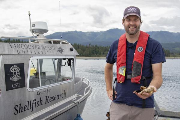 Dr. Timothy Green, VIU’s Canada Research Chair in Shellfish Health and Genomics stands on a dock next to a boat holding oysters in his hand.