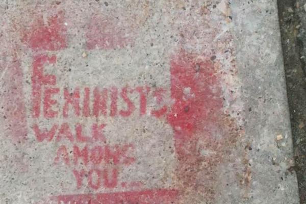 A light grey concrete sidewalk is spray painted with the phrase "Feminists Walk Among You."