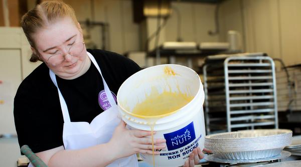 Faithe Haley, wearing a black shirt and white apron, pours sugar into pies in an industrial kitchen.