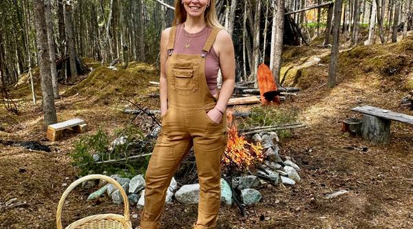 A woman wearing tan overalls is standing in a clearing in the forest in front of a campfire. 