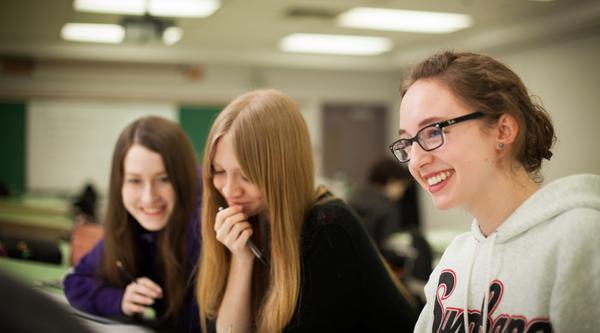 three female students in a classroom smiling and looking at an assignment