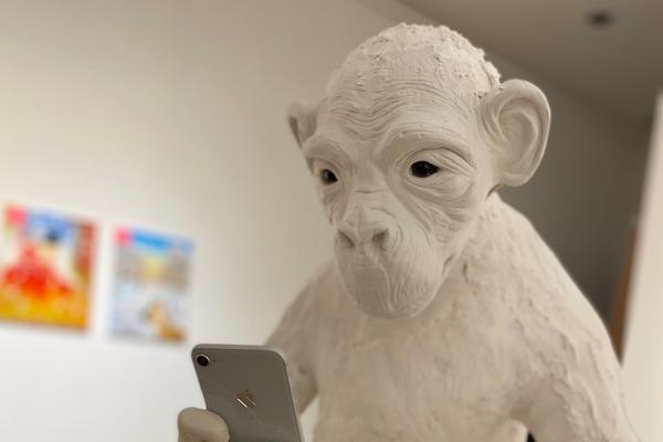 An ape made of white plaster and clay holds a cell phone, which it stares at intensely