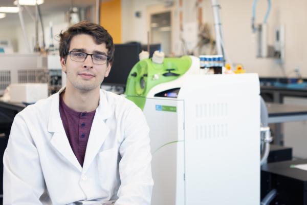 Joseph Monaghan sits in a VIU Chemistry lab, wearing a white lab coat.