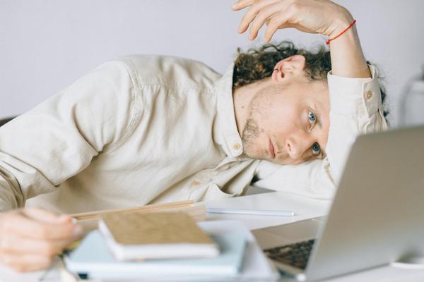 Student leaning on his arm staring dejectedly at a laptop screen with a pile of books beside him