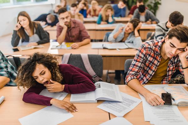 a student on the left yawns while the student next to her has his hand on his face while in a classroom with lots of students in the background