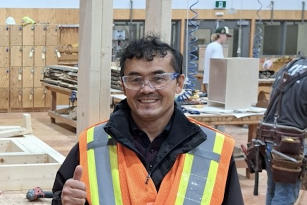 Ezat Haidary wering a reflective vests and safety goggles in the carpentry shop, giving a thumbs up and smiling at the camera.