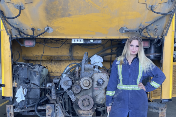 Madeline kozubal wearing mechanics coveralls standing in front of a school bus engine and smiling at the camera