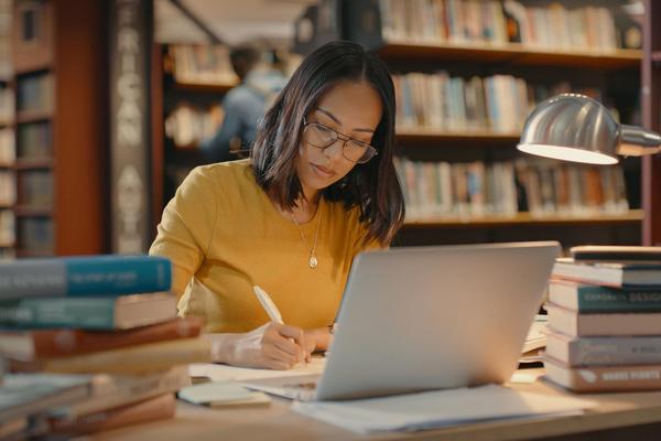 A woman sits at a desk in a library with a laptop open surrounded by books