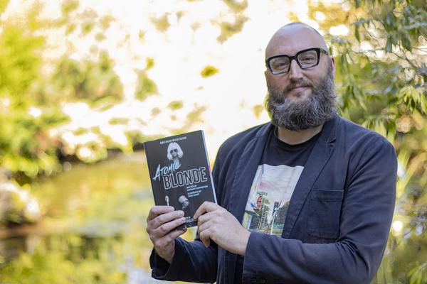 Dr. Theo Finigan, a VIU English Professor, holds up the book Atomic Blonde while standing in front of the VIU koi pond.