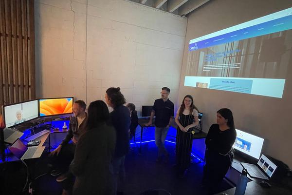 Students gather around three screens on a desk with a projection of an image on the wall behind them.