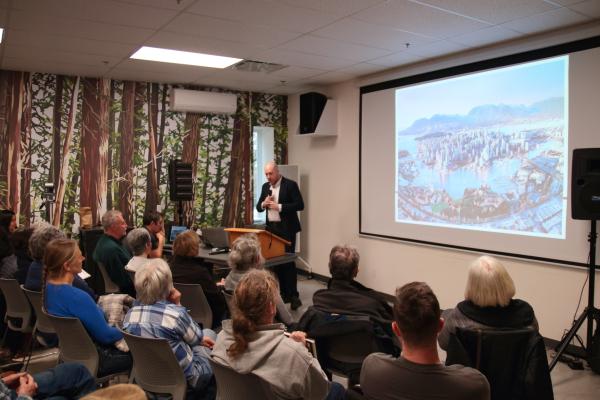 a man speaks next to a screen in front of a packed room