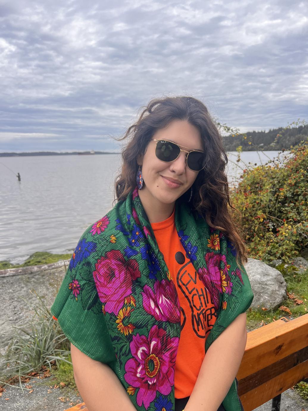 Alyona stands near a lake wearing a green shawl with pink, blue and yellow flowers over an Every Child Matters shirt.