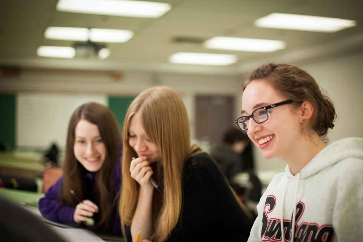 three female students in a classroom smiling and looking at an assignment