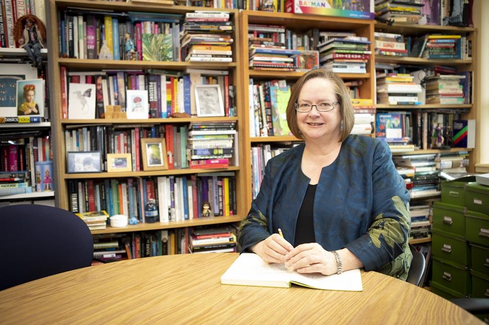 A female professor sits at a desk in an office with books and papers in the background