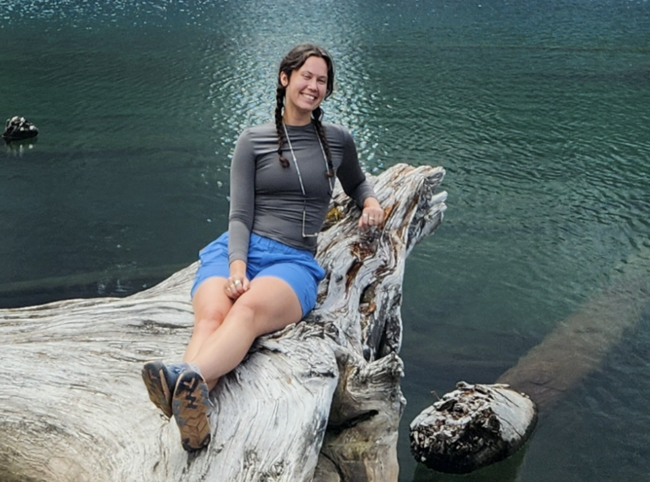 Holly Faithfull sitting on a log surrounded by a lake and smiling at the camera.