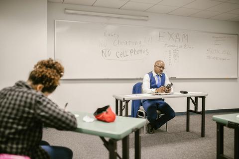 An instructor sits at the front of a classroom adjudicating an exam, one student is seen writing a test
