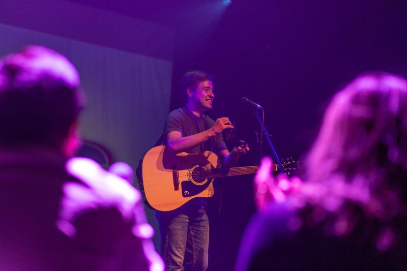 Man with guitar on stage with backs of members of crowd visible