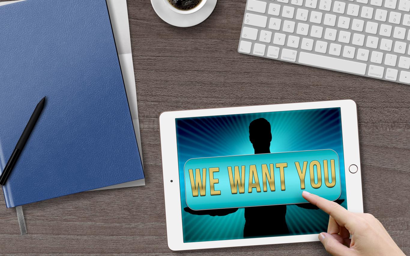keyboard, ipad that says We Want You, folders, pens and cup of coffee on a desk